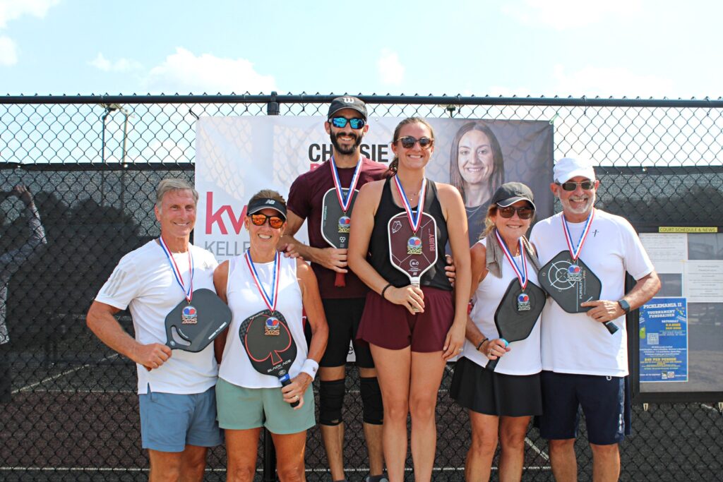 Six adults stand on a pickleball court, smiling and holding paddles and medals. Three are on a higher podium step. They pose in front of a chain-link fence with a large banner and a schedule posted.