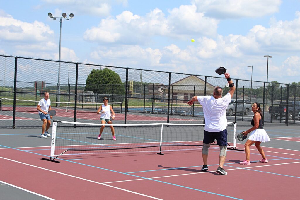 Four adults play pickleball on an outdoor court. Two men and two women are mid-game, with one man leaping to hit the ball. The court is surrounded by a chain-link fence on a sunny day.