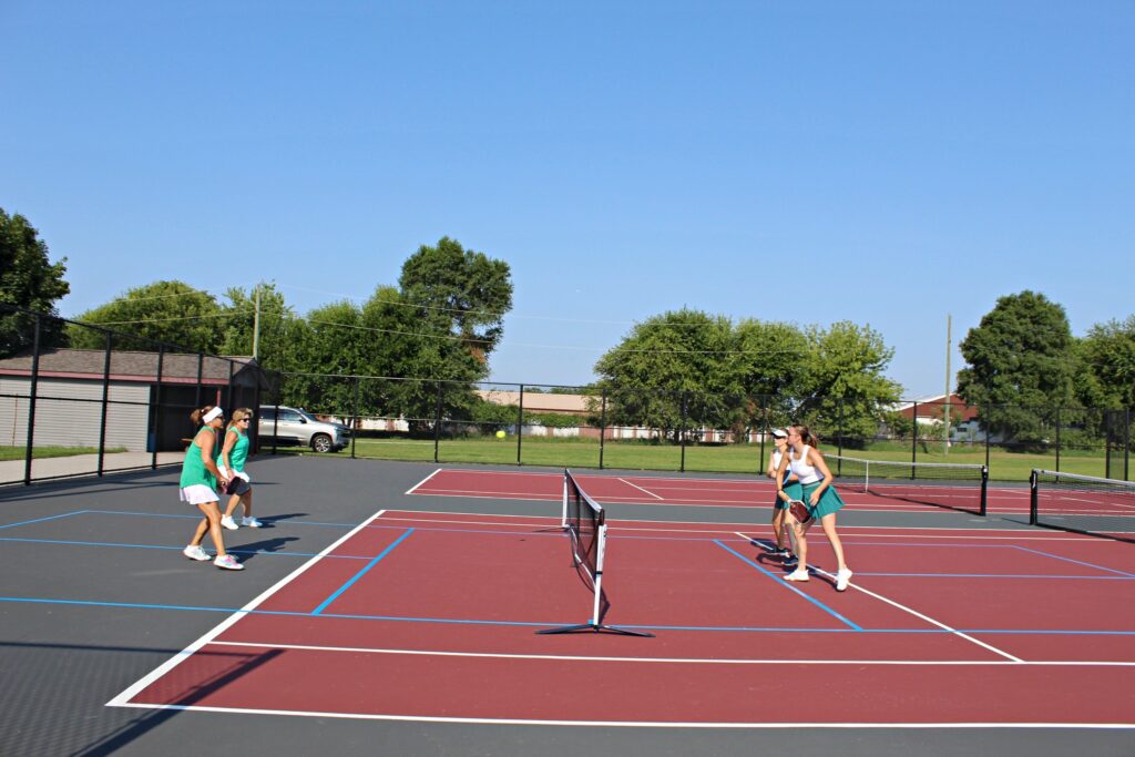 Four women play a doubles game of pickleball on outdoor red and blue courts, surrounded by a black fence, with trees and buildings visible in the background under a clear blue sky.
