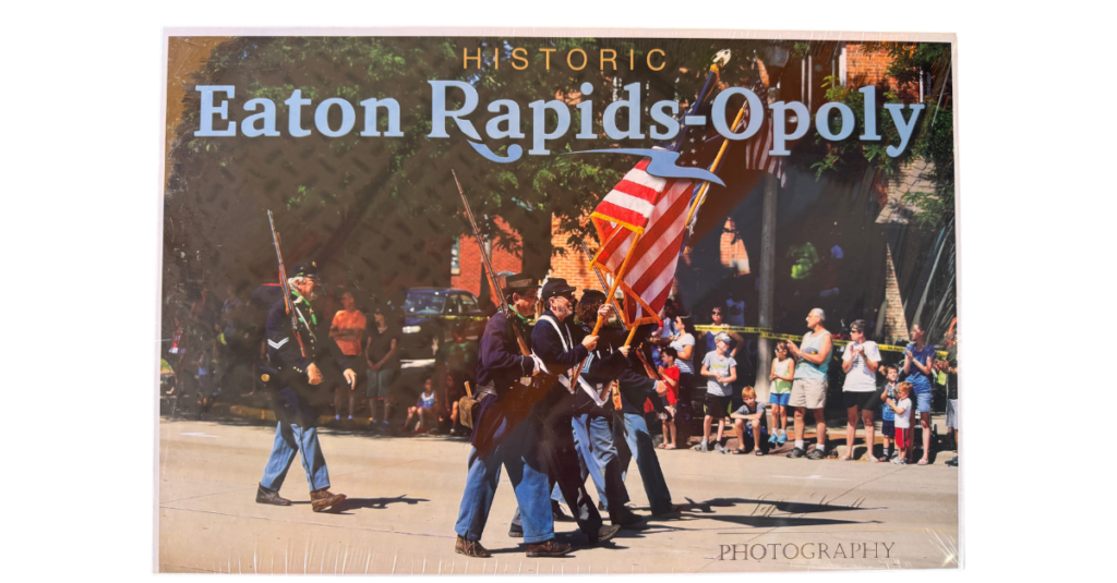 A group of people in historic military uniforms march with American flags in a parade, as a crowd watches from the sidewalk. Text above reads, Historic Eaton Rapids-Opoly.