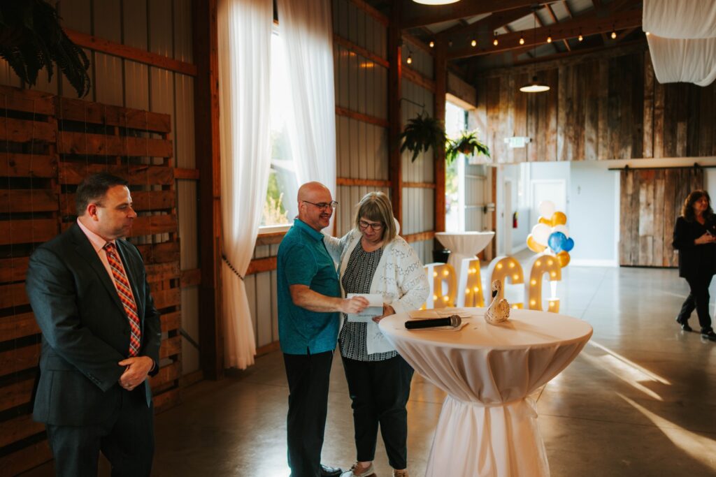 Three people are at an indoor event with rustic decor. A man and woman stand together smiling by a round table with papers. Another man in a suit stands nearby. RACC letters and balloons are in the background.
