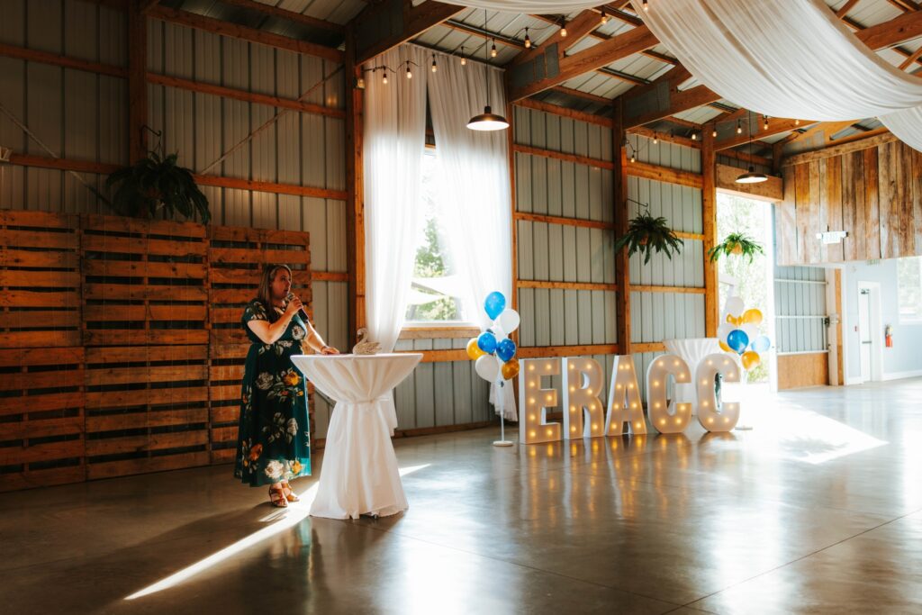A woman in a floral dress stands at a tall round table in a decorated event space with wood and metal walls. Large light-up letters spell “GRAD,” and blue, white, and yellow balloons are nearby. Draped fabric and string lights hang above.
