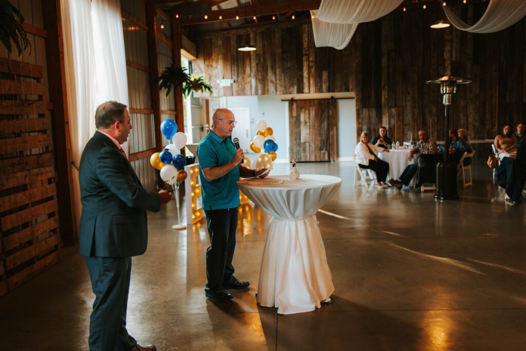 Two men stand at a cocktail table with a microphone at an indoor event. Guests sit at tables in the background, and white and blue balloons are arranged near a wooden wall. The room has rustic decor and soft lighting.