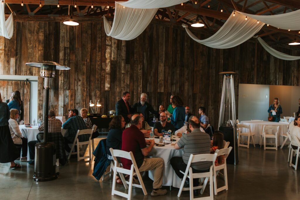 A group of people sit at round tables covered with white cloths in a rustic indoor event space with wooden walls and draped fabric on the ceiling. Some people are standing and talking, while others are seated and eating.