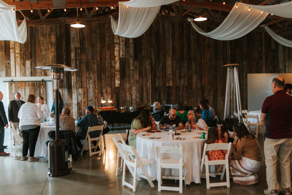 A group of people sit and stand around round tables with white tablecloths in a rustic indoor venue with wood-paneled walls, draped white fabric on the ceiling, and heaters. Some guests are talking or eating.