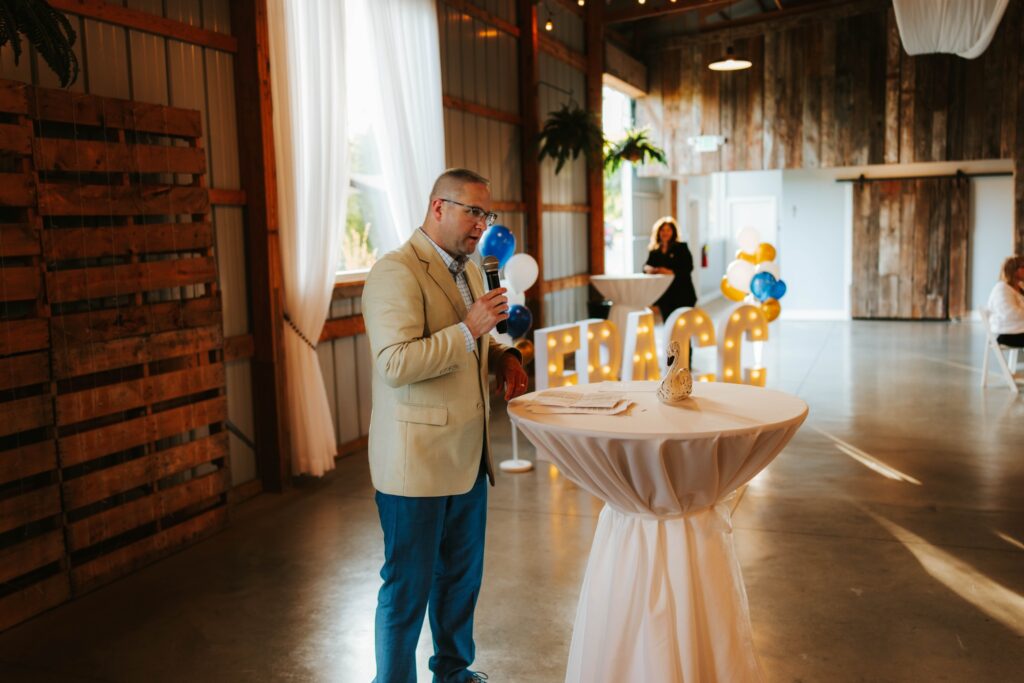 A man in a tan blazer speaks into a microphone at a round cocktail table in a rustic event space, with large lit-up letters and balloons in the background.