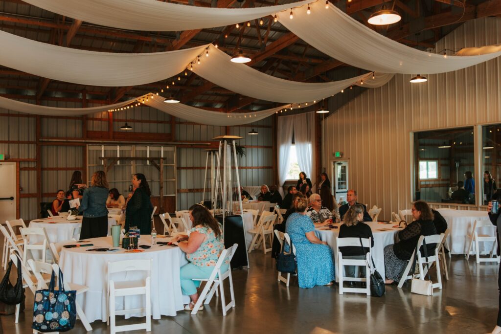 People sit and talk at round tables covered with white tablecloths in a spacious, rustic event hall with draped white fabric and string lights overhead. Some guests stand and mingle while others are seated and engaged in conversation.