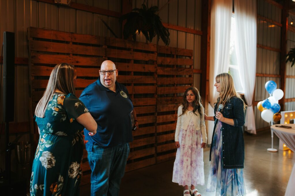 Four people standing and conversing indoors near a wooden backdrop; two women in floral dresses, a man in a navy polo shirt, and a young girl in a white and pink dress. Balloons and sunlight through windows are visible in the background.