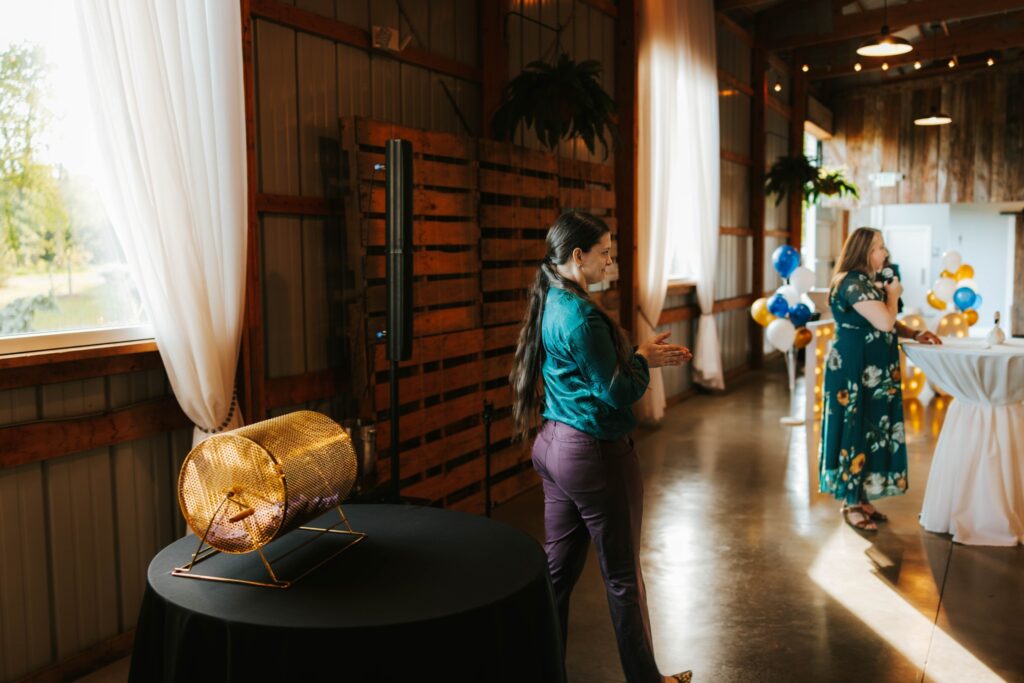 A woman in a teal blouse stands near a gold raffle drum on a black table at an indoor event with balloons and tall white curtains; another woman in a floral dress stands in the background.