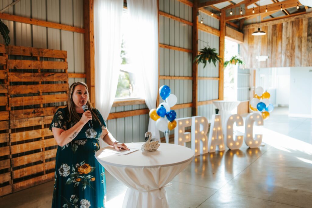 A woman in a floral dress speaks into a microphone at a round table with a swan decoration. Behind her are balloons and large marquee letters spelling FRACC in a bright, rustic event space.
