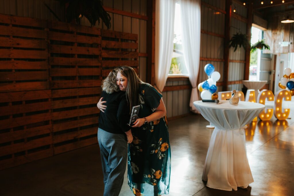 Two women hug in a warmly lit event space with wooden walls, large windows, and decorative balloons. One woman wears a floral dress and holds a plaque. A round table with a white cloth stands nearby.