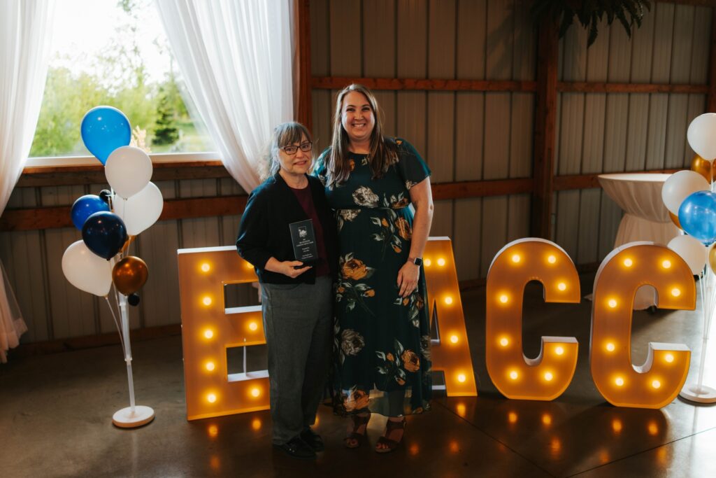 Two women stand smiling indoors near lit marquee letters reading EACC. One woman holds an award. There are balloons on each side and white curtains with outdoor light coming through the window behind them.