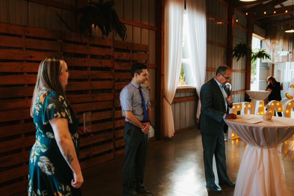 Three people stand in a decorated indoor venue, with one man in a suit speaking into a microphone at a tall round table. Warm lighting and sheer curtains create a festive atmosphere.