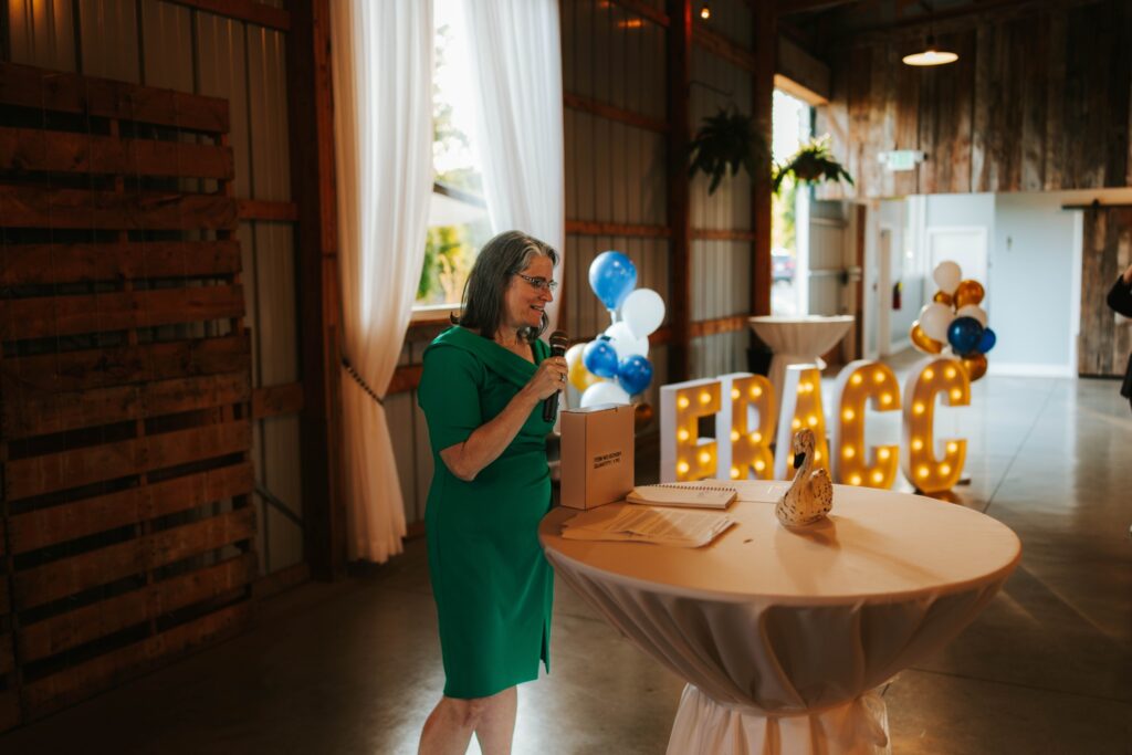 A woman in a green dress stands at a round table with a microphone, speaking at an indoor event. Behind her are lit-up letters spelling FRACC and blue, white, and gold balloons.