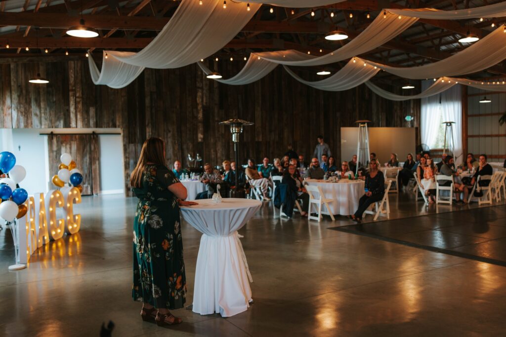 A woman in a floral dress stands at a round table, speaking to a seated audience at an indoor event decorated with draped fabric, string lights, and blue and gold balloons.