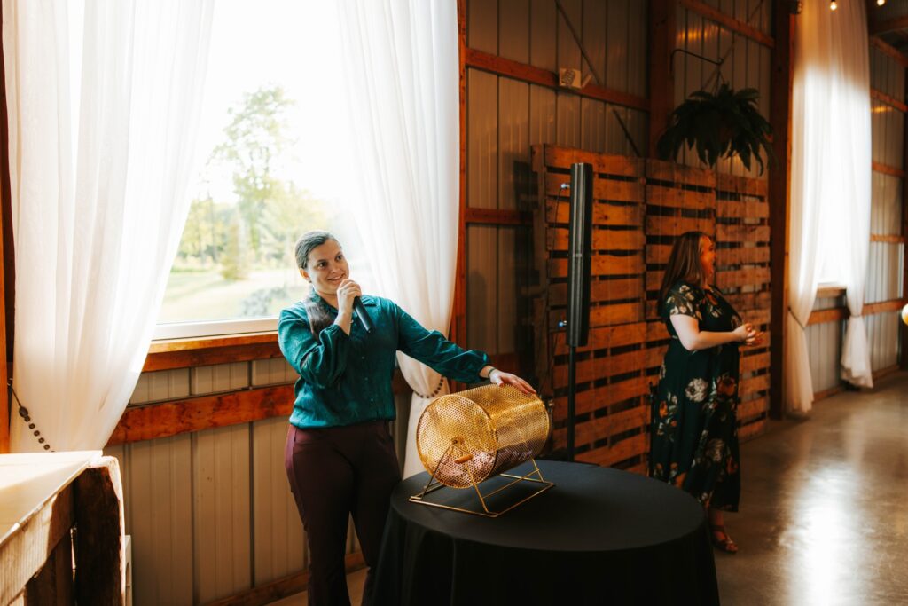 A person stands next to a raffle drum on a table, speaking into a microphone. Bright natural light streams through a window with white curtains in a rustic venue. Another person stands in the background near wood paneling.
