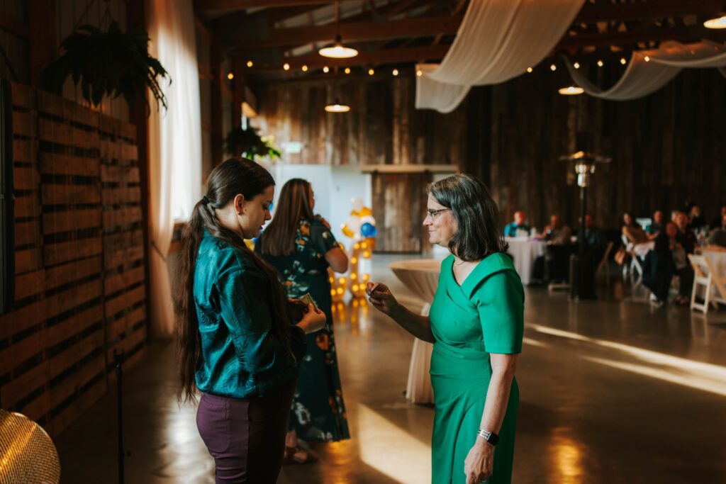 Two women stand and talk in a warmly lit event space with wooden walls and draped fabric on the ceiling. One woman wears a green dress, the other a teal blouse and dark pants. Guests and round tables are visible in the background.