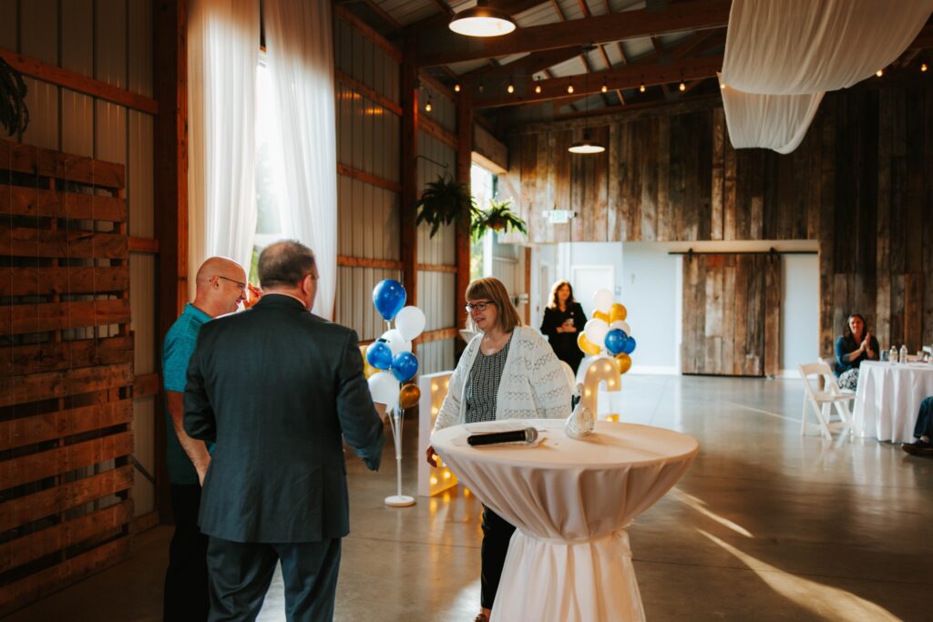 A woman stands at a cocktail table, talking to two men in a rustic event space decorated with blue and white balloons and string lights. Other people sit at round tables in the background.