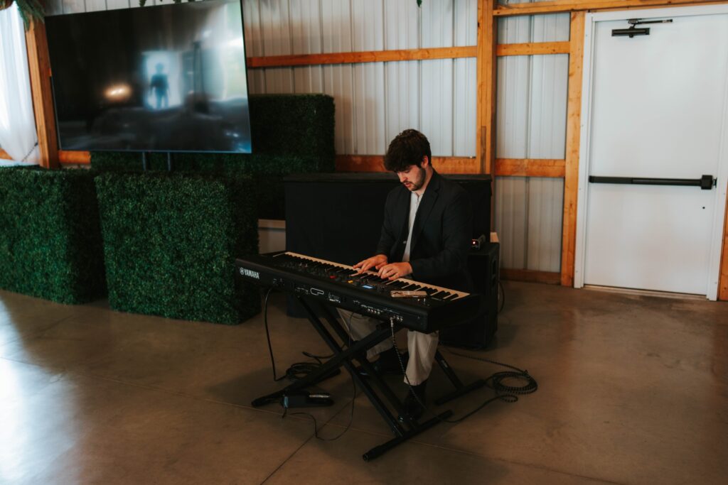 A man in a suit jacket sits and plays a keyboard inside a room with concrete floors, artificial greenery, a large TV, and a closed white door in the background.