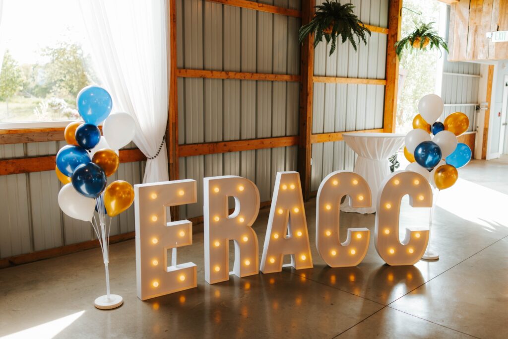 Large illuminated letters spelling ERACC are displayed indoors, flanked by clusters of blue, gold, and white balloons. The setting features wooden beams, a white curtain, and natural light coming through windows.