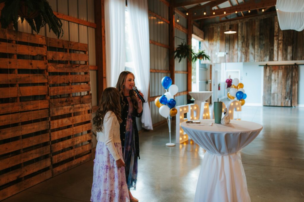 Two women in floral dresses stand near a cocktail table in a rustic event space with wood walls, string lights, and colorful balloons. One woman speaks into a microphone.