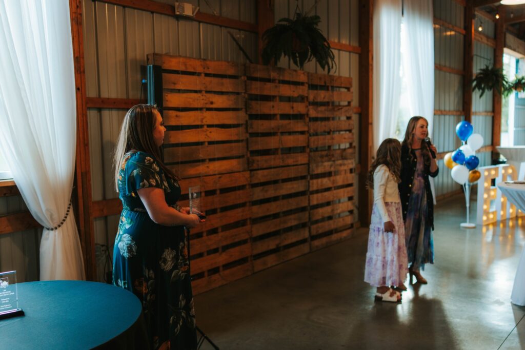 Three women stand in a decorated event space with wooden walls, white curtains, and balloon arrangements; two are speaking into microphones while one stands to the side holding a glass plaque.