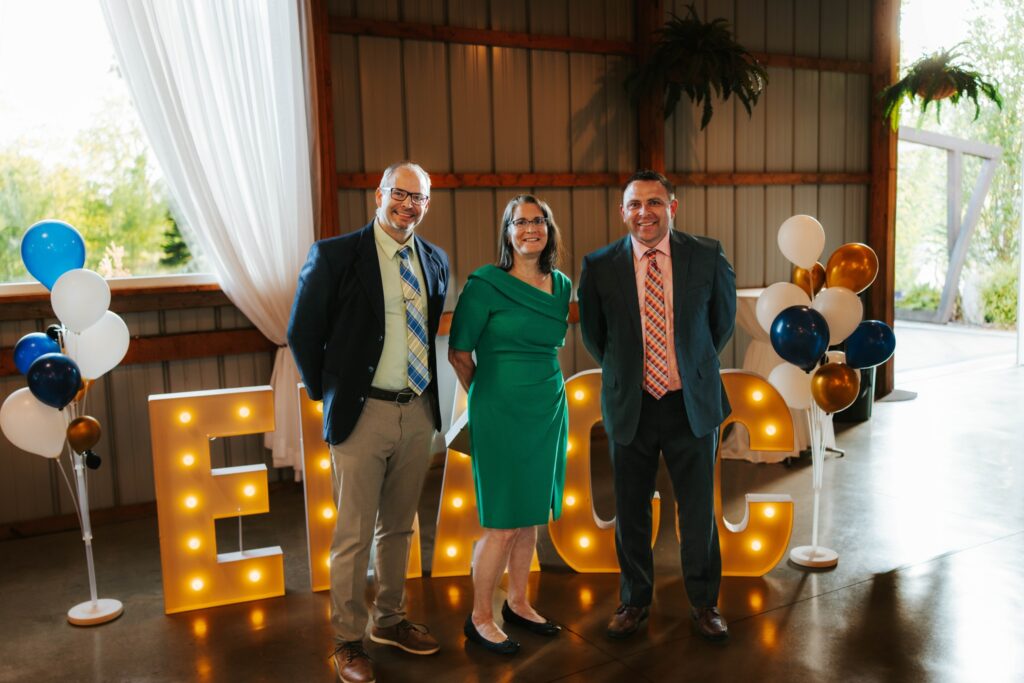 Three people in formal attire stand smiling indoors in front of large, lit-up letters reading EMCC, with decorative balloons and natural light coming from windows in the background.