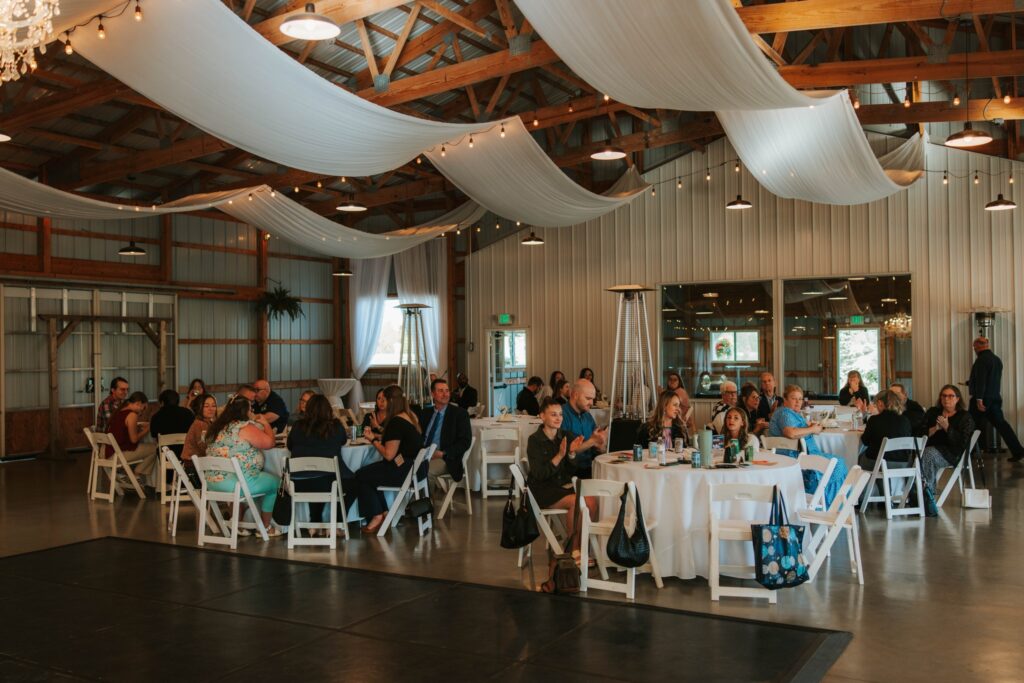 Guests seated at round tables in a decorated barn-style venue with draped white fabric and string lights across the ceiling, attending an event and facing the front, some clapping, and others engaged in conversation.