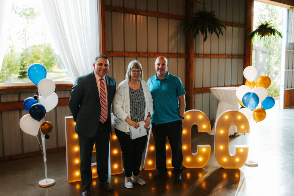 Three people stand smiling indoors next to gold and white balloon arrangements and large lit-up MVCC letters. The setting has wood and metal walls, with sunlight coming through a window and an open door.