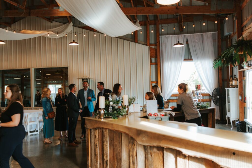 A group of people in formal attire socialize near a rustic wooden bar inside a decorated barn with string lights, draped white fabric, and large windows letting in natural light.
