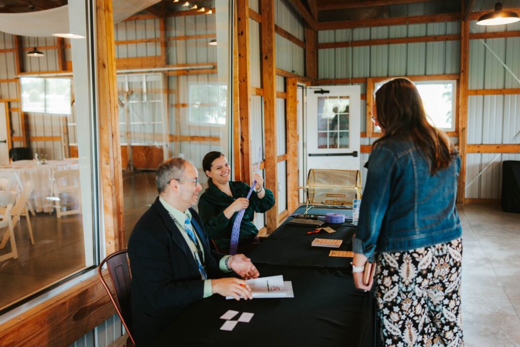 Two people sit at a table handing out tickets or name tags to a woman standing in front of them, inside a rustic venue with wood beams and large windows. The atmosphere appears welcoming and organized.