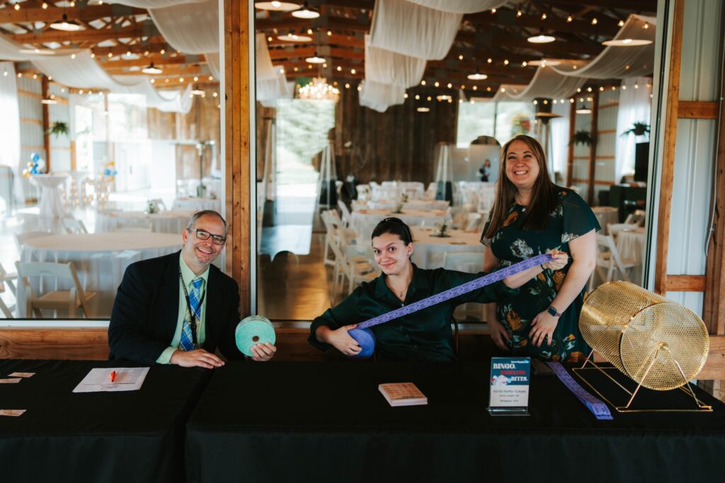 Three people sit and stand behind a black table at an event check-in area. One holds a roll of tickets, another holds a large tape measure, and a third stands smiling. A raffle drum is on the table, and decorated tables are in the background.