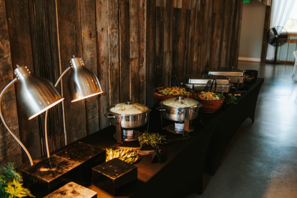 A buffet table set against a rustic wooden wall features covered chafing dishes, large salad bowls, and two silver heat lamps, ready for a catered meal in a spacious, well-lit room.