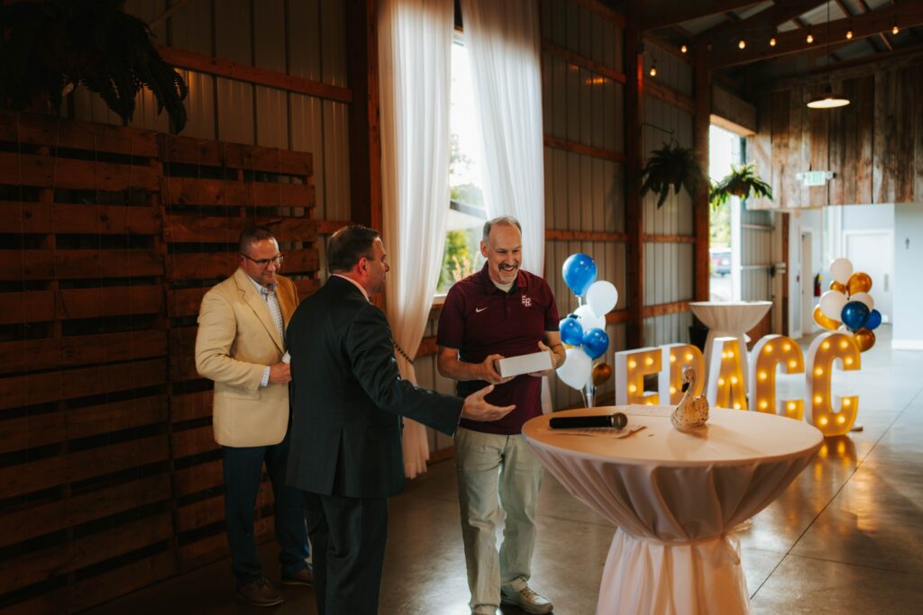 Three men stand near a table at an indoor event. One man in a maroon shirt is smiling and holding a box, while two others in suits look on. Balloons and large, lit “FRACC” letters are visible in the background.