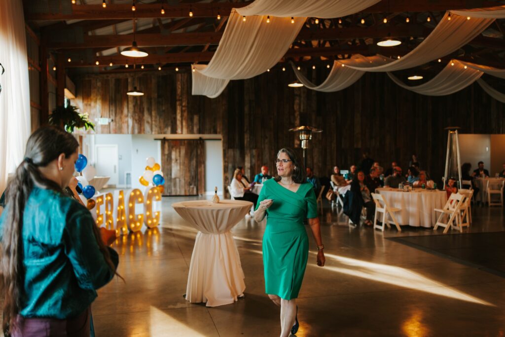 A woman in a green dress walks past a cocktail table at a party in a decorated barn. Guests are seated at round tables, and large “PEACE” letters with balloons are in the background. Warm light fills the room.