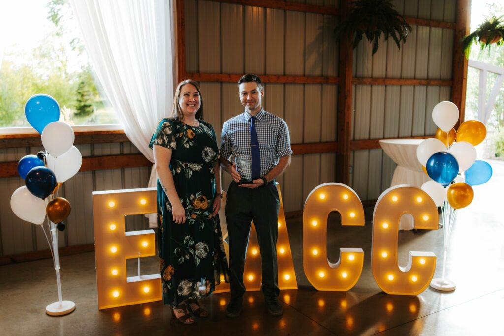 A woman and a man stand smiling indoors next to large illuminated letters reading EMCC. They are dressed formally, with balloons and cocktail tables in the background, suggesting a celebratory event.