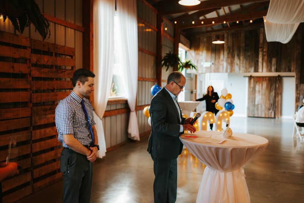Two men stand at a tall round table in a decorated event space with balloons and string lights. One man studies papers while the other stands nearby, both wearing business attire. The atmosphere is formal yet festive.