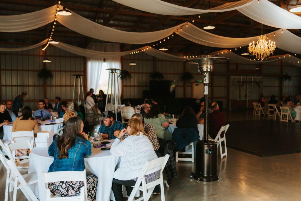 People sit at round tables covered with white tablecloths in a large indoor venue decorated with draped fabric, string lights, and a chandelier. The atmosphere appears relaxed and social.