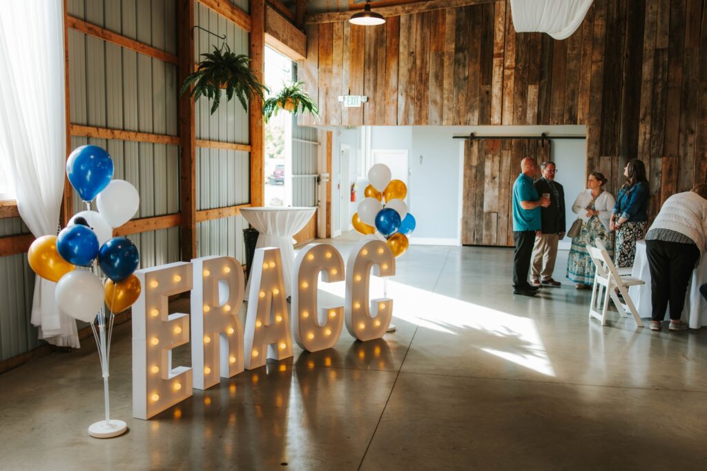 Large light-up letters spell ERACC beside a cluster of blue, white, and gold balloons in a rustic event hall with wooden walls. Several people stand and chat near white chairs in the background.