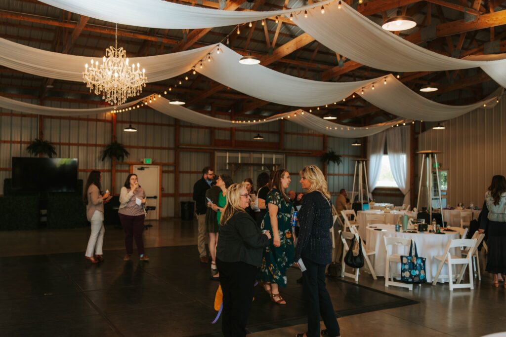 A group of people gather and converse in a decorated event space with draped white fabric, a chandelier, round tables, and wooden beams overhead. Some stand on a dance floor while a few others talk in the background.