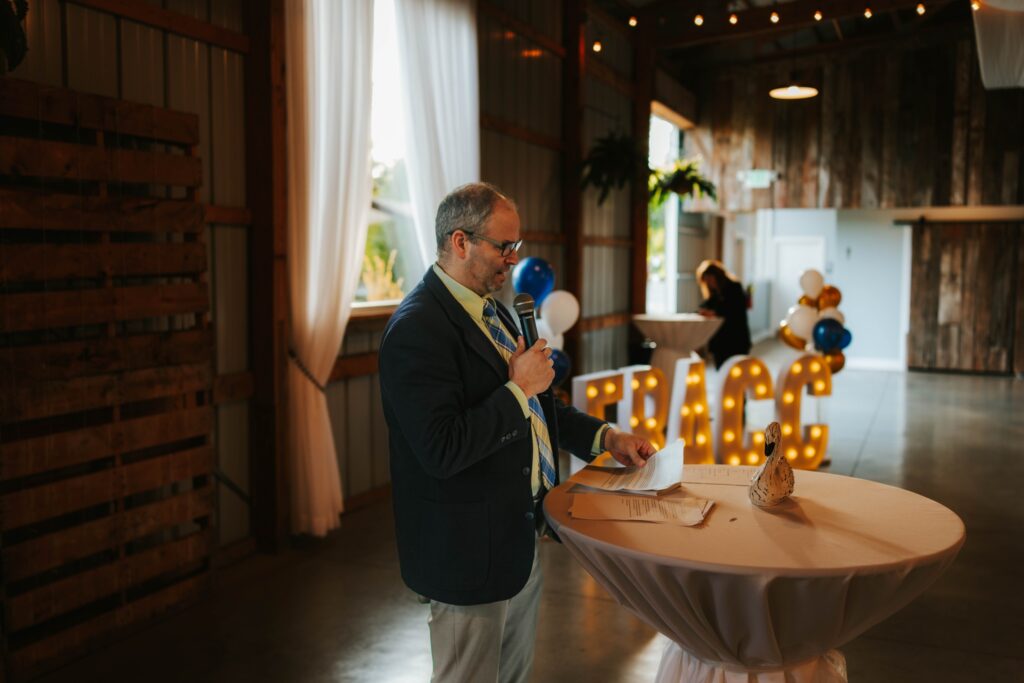 A man in a suit speaks into a microphone at a round table with papers and a hat on it. Behind him are large, illuminated letters spelling “GRACE,” white and gold balloons, and rustic decor in a softly lit event space.