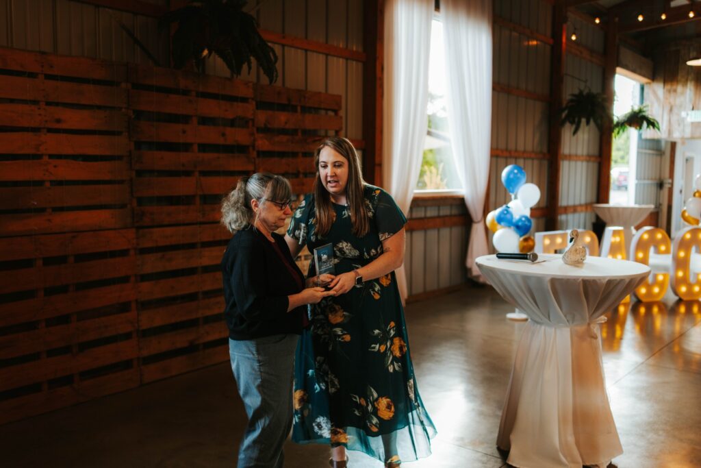 Two women stand and talk inside a decorated event space with wooden walls, string lights, blue and white balloons, and a round table with a white tablecloth in the background.