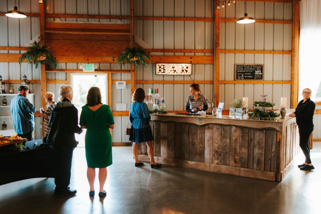 Several people stand and wait in line at a rustic wooden bar inside a spacious, well-lit venue with high ceilings, plants, and a glowing BAR sign on the wall above the counter.