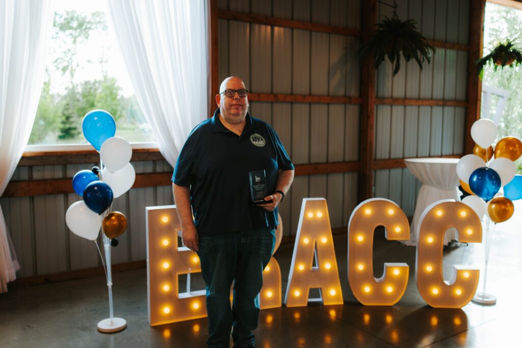 A man holding an award stands in front of large light-up letters spelling BRACC, with white, blue, and gold balloons on either side, inside a decorated venue with natural light from a window.