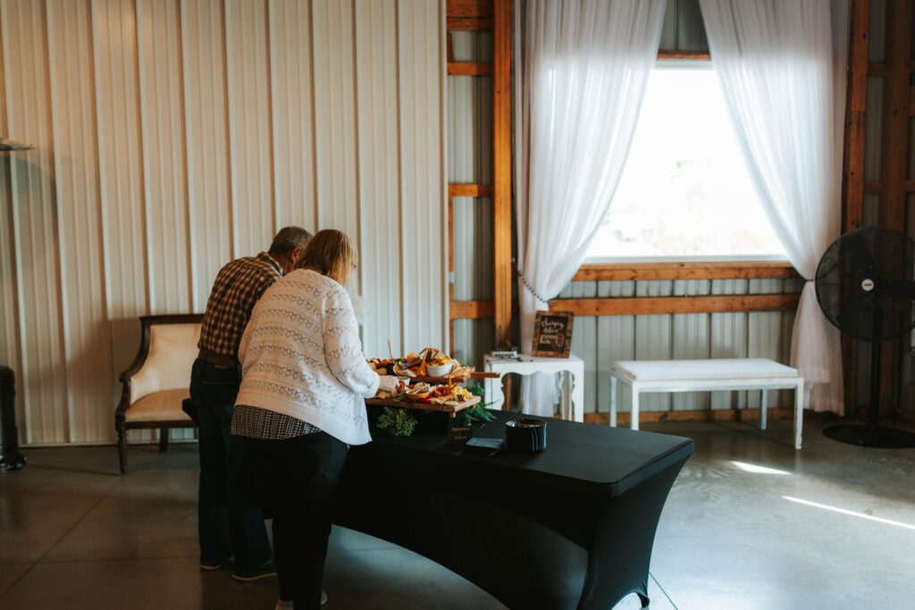 Two people stand at a black table with an assortment of food inside a bright room with metal walls, a white curtain, a bench, a fan, and a chair in the background.