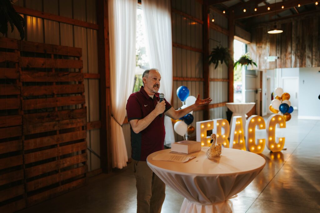 A man speaks into a microphone at a round table in a decorated indoor venue, with lighted FRACC letters and balloons in the background.