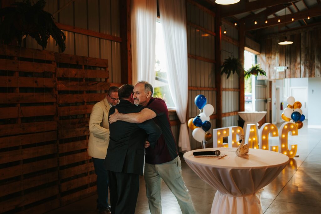 Two men hug in a warmly lit event space next to a round table and large illuminated letters spelling FRACC. Balloons and a microphone are visible, with a third man standing nearby, smiling.