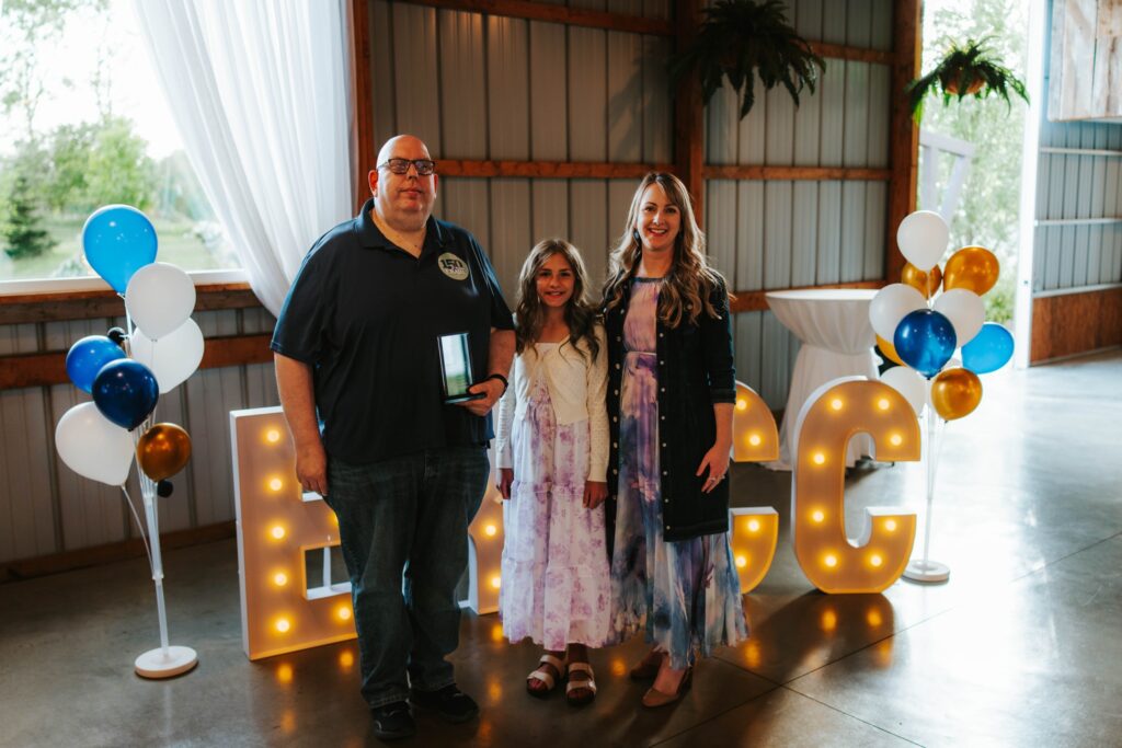 A man, a young girl, and a woman stand smiling in front of large illuminated ECC letters, with balloon decorations on each side, inside a decorated event space with natural light.