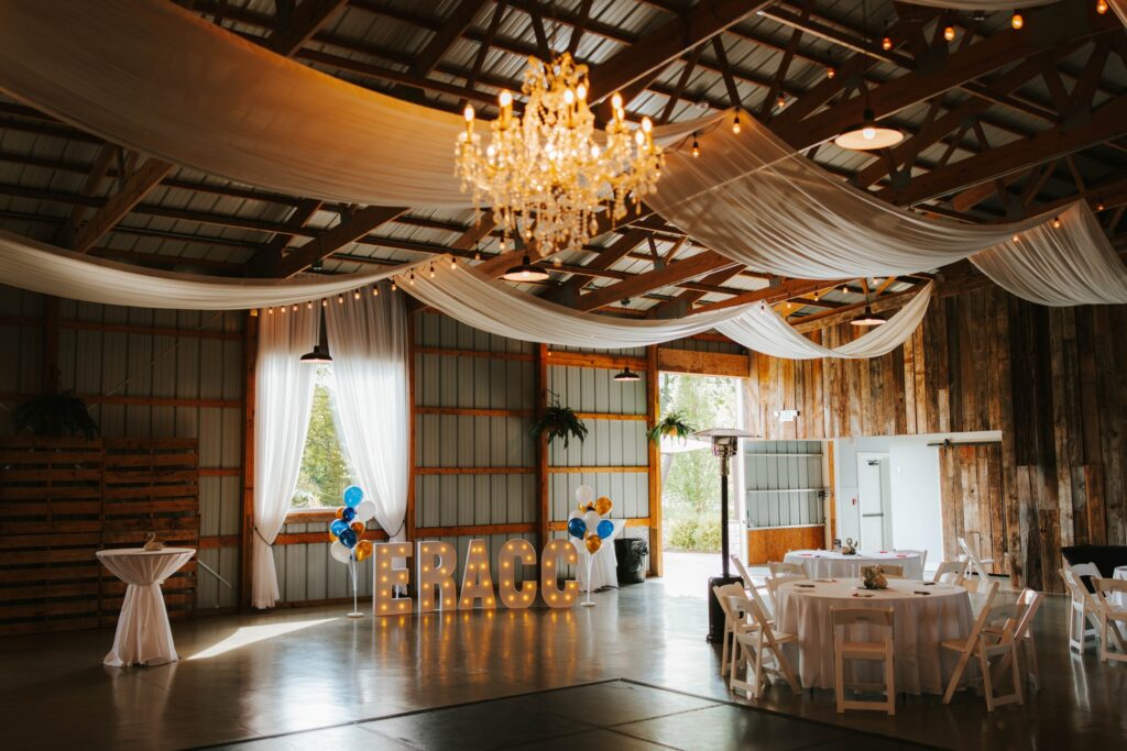 A rustic event space with wooden walls, draped white fabric ceiling, chandelier, round tables with white chairs, and large lit-up letters spelling FRACC with colorful balloons nearby.